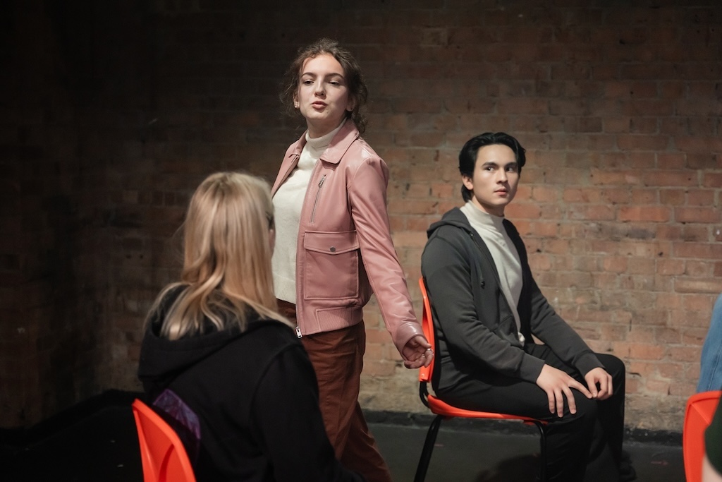 Action shot of the spring 2024 Shakespeare in Performance at RADA final presentation of Julius Caesar. An actor wearing a light pink coat stands in mid-performance, as two other actors, seated in bright orange chairs, look on.